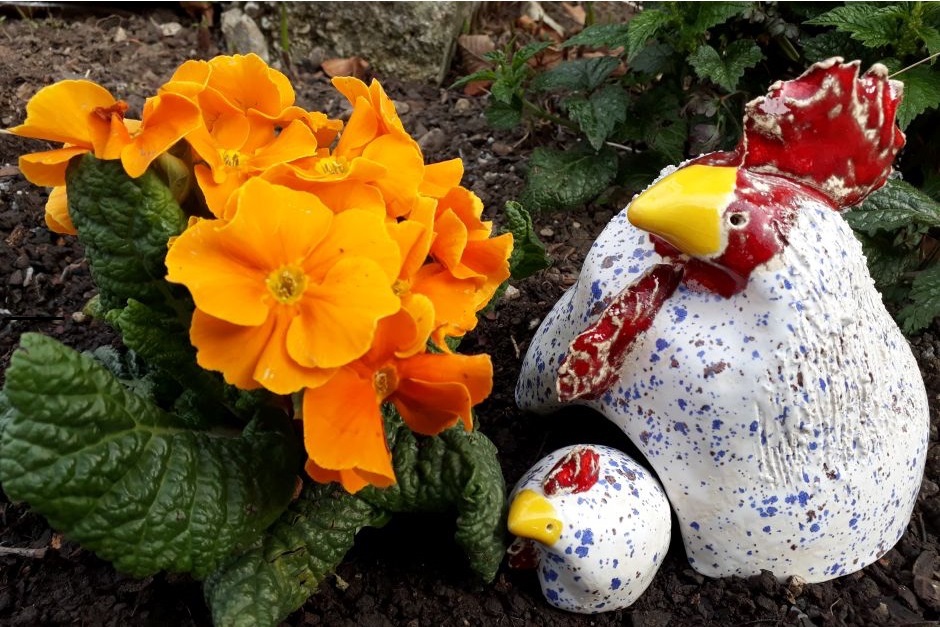 Bild mit oranger Blume und einem Mutterhuhn aus Keramik und einem Babyhuhn daneben, in grau mit roter Schleife.